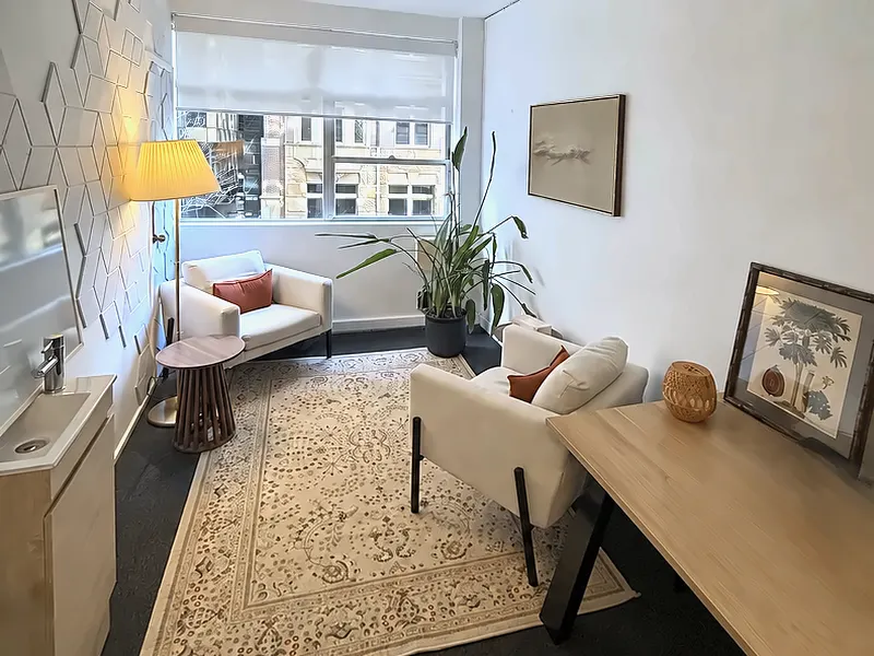 Psychology consulting room at Sydney Sleep & Insomnia: two cream armchairs on a patterned rug, a floor lamp, a plant and a large window, with a desk along the right-hand wall.